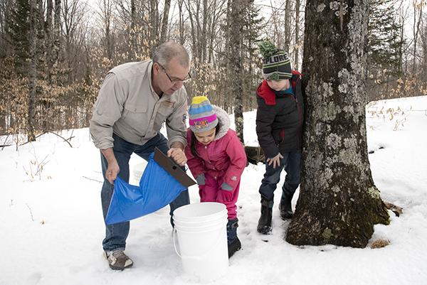 Pete Kolbas shows 4-H members, Isabella Boyear and Aiden Cronkright how to empty the sap after tapping the maple tree. The youth enjoyed learning the process at Old Maki Jawbone Ranch located in Iron County.