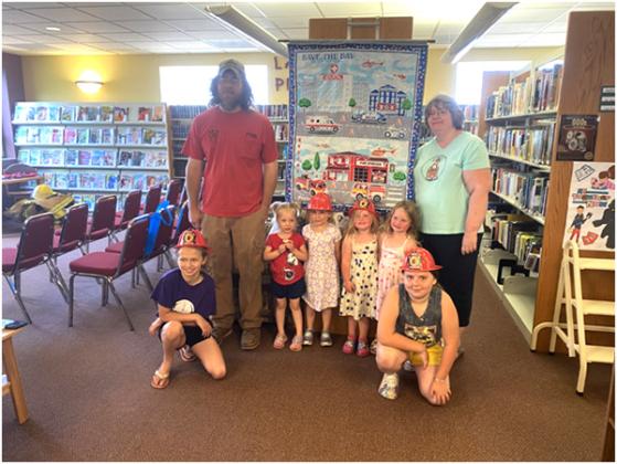 Peyton Jackson, Fire Chief Fabbri, Ivy Skogman, Charlotte Rodriguez, Rory Alquist, Revy Alquist, Library Director Evelyn Gathu and Audrey Rodriguez standing by a Community Heros Quilt that Library Clerk Charlene Bendick made and gave to be put up at City Hall.
